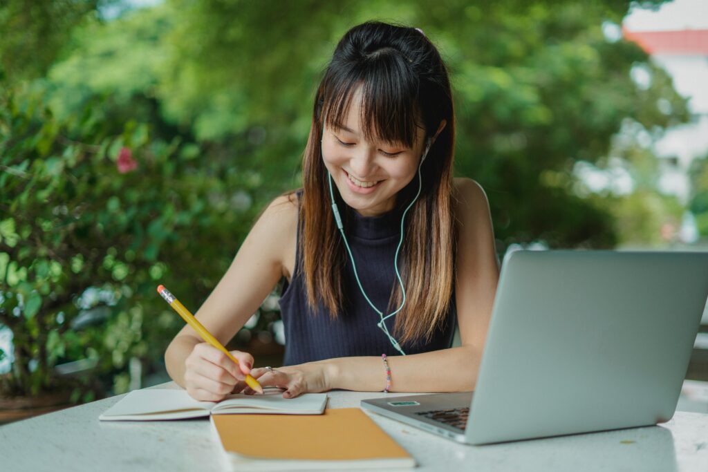 Positive young Asian female student with earphones writing in copybook while doing homework at table with laptop in street cafeteria